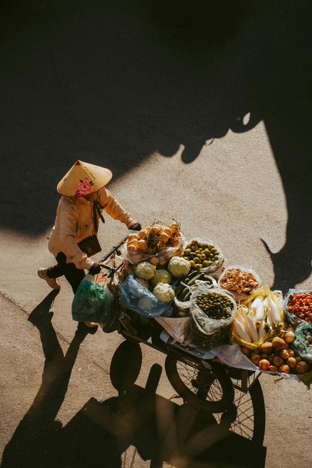 Street vendor pushing a bicycle cart loaded with various fruits and vegetables under warm sunlight in Hanoi, Vietnam.